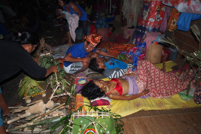 Wooden floor covered with cloth, blankets, thin mattresses; in the right part of the picture, two people of female appearance lying beneath each other on the floor lower body covered with blanket; a shaman crouches at the head of the one person and seems to talk to the person; the shaman places a plant on the person's stomach; on the left side of the picture another person holds a house made of plants for the spirit; a wooden doll lies on the floor; in the background many people watching; colorful cloth