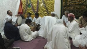 Apparently a group men sitting on the floor, some of them clapping, some playing drums; except form two they are dressed in long white dresses, some wearing a kufi (head wrap)