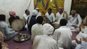 Apparently a group men sitting on the floor, some of them clapping, some playing drums; except form two they are dressed in long white dresses, some wearing a kufi (head wrap)