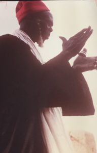 Person in black cloth with white shawl, red headwear and beard raising the hands in front the chest, facing write