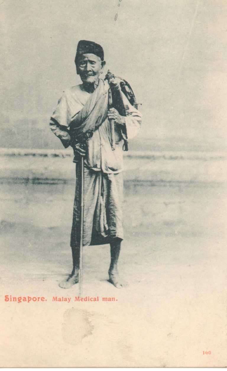 The picture shows a postcard which depicts a Malay medicine man standing and holding a stick.